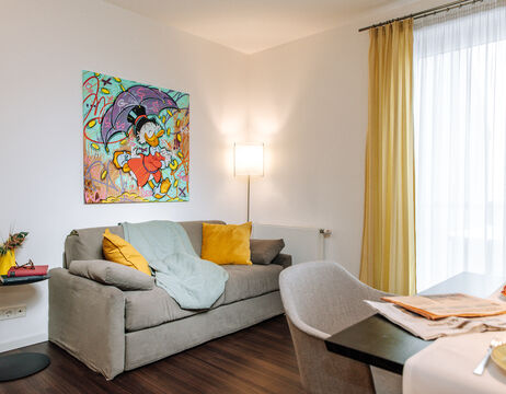 Ein gemütliches Wohnzimmer mit einem grauen Sofa, gelben Kissen, farbenfroher Wandkunst und gelben Vorhängen.