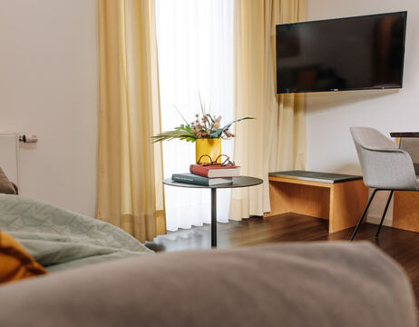 Modernes Wohnzimmer mit einem kleinen runden Tisch, Büchern, Gläsern, Blumen, Fernseher, Stühlen und gelben Vorhängen.