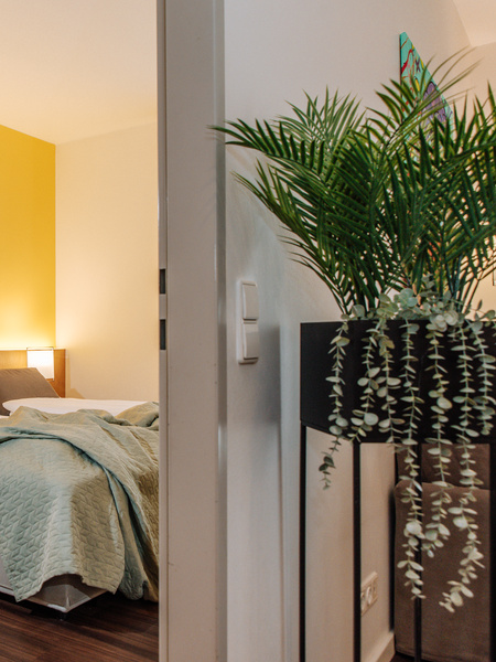 A cozy bedroom with a yellow accent wall, a double bed, a green blanket and a potted plant in the foreground.