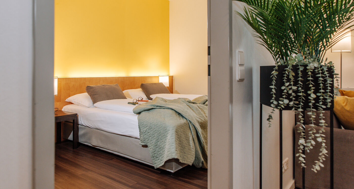 Ein gemütliches Schlafzimmer mit einer gelben Akzentwand, einem Doppelbett, einer grünen Decke und einer Topfpflanze im Vordergrund.