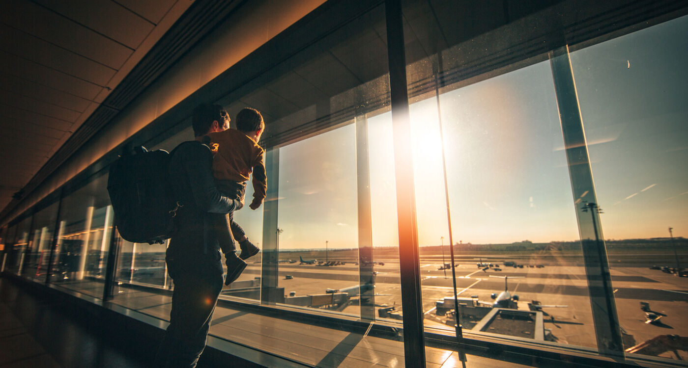 Ein Erwachsener, der ein Kind im Arm hält, schaut aus dem Flughafenfenster auf Flugzeuge bei Sonnenaufgang oder Sonnenuntergang.